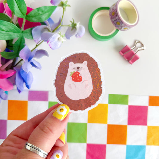 A glittery sticker of a small cute hedgehog holding a little strawberry in her hands.