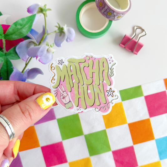 A holographic sticker with text saying 'Matcha Hun' in funky bubble writing and other hand illustrated elements around the text. The colours are pink and green.