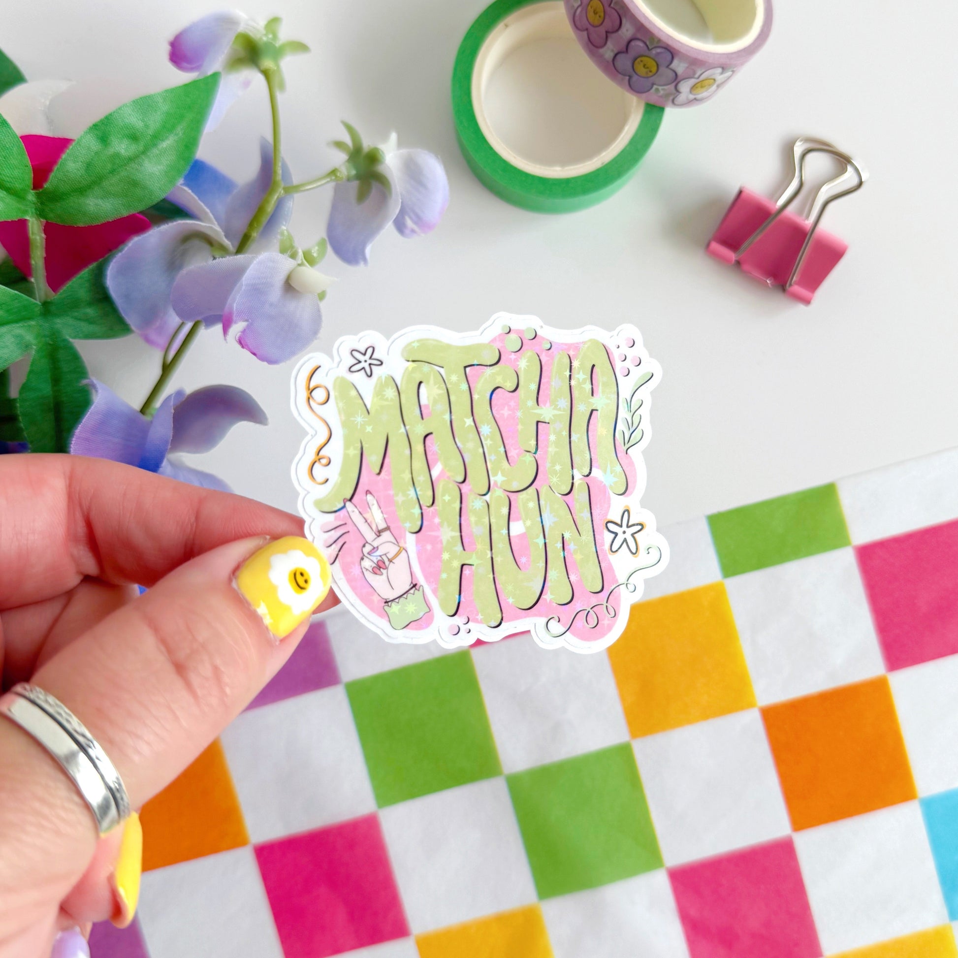 A holographic sticker with text saying 'Matcha Hun' in funky bubble writing and other hand illustrated elements around the text. The colours are pink and green.