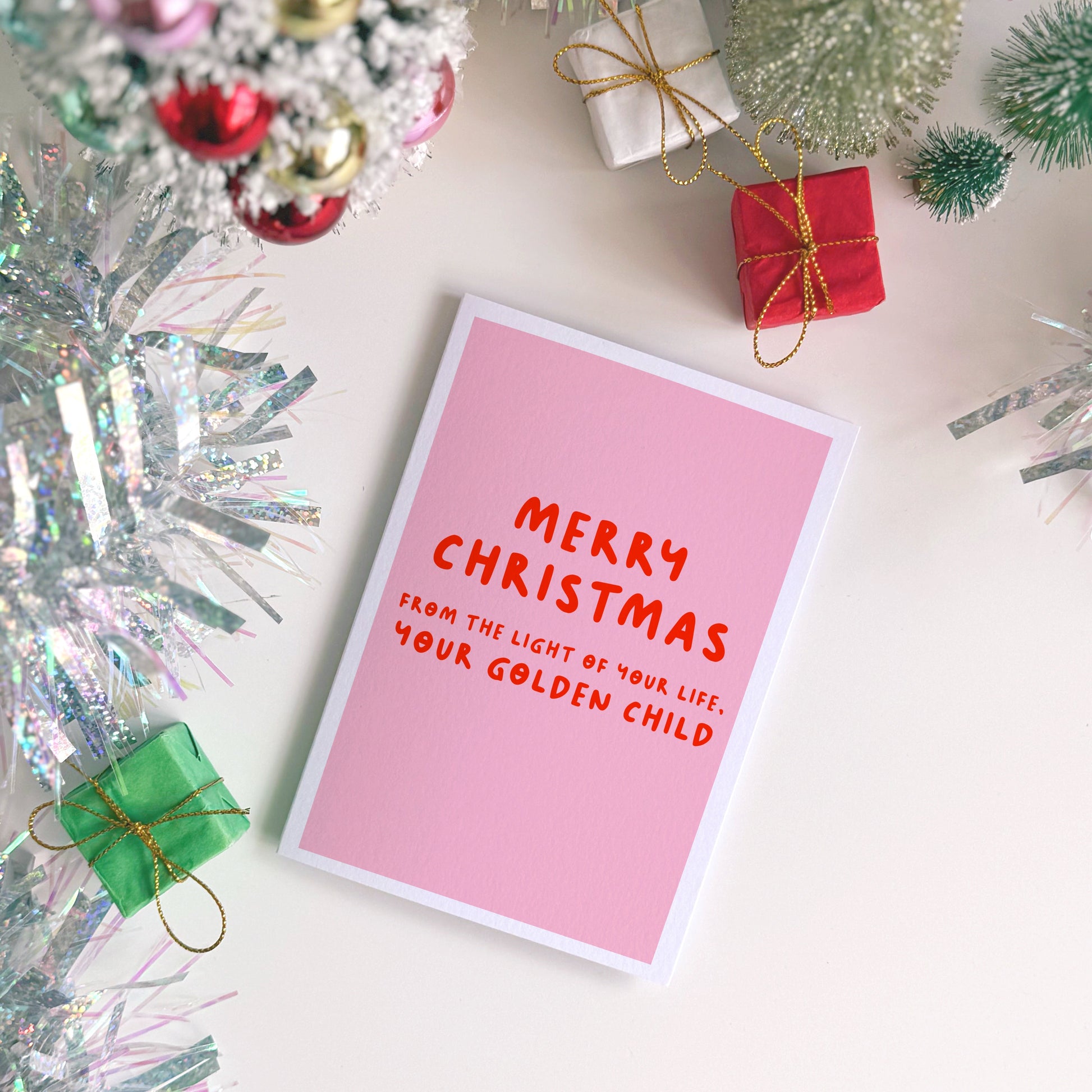 Pink Christmas card with red text on a white surface with festive decorations.