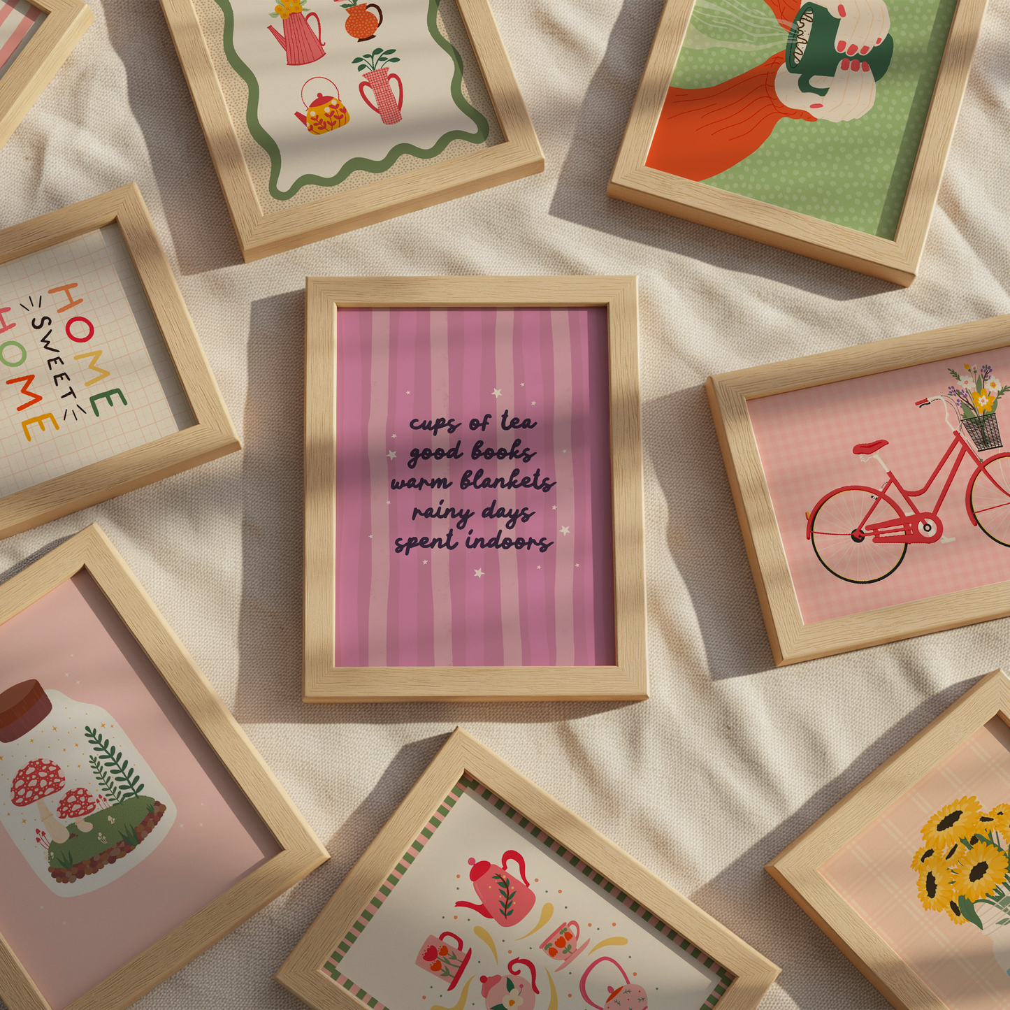 A styled flat-lay of multiple small light wood picture frames arranged on a cream fabric background. The central frame contains pink vertical stripes with black handwritten text reading, 'cups of tea good books warm blankets rainy days spent indoors.