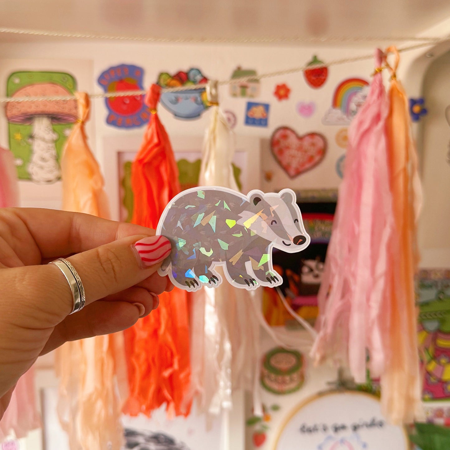 Hand holding a cute badger sticker in front of a shelf with toys and decorations.