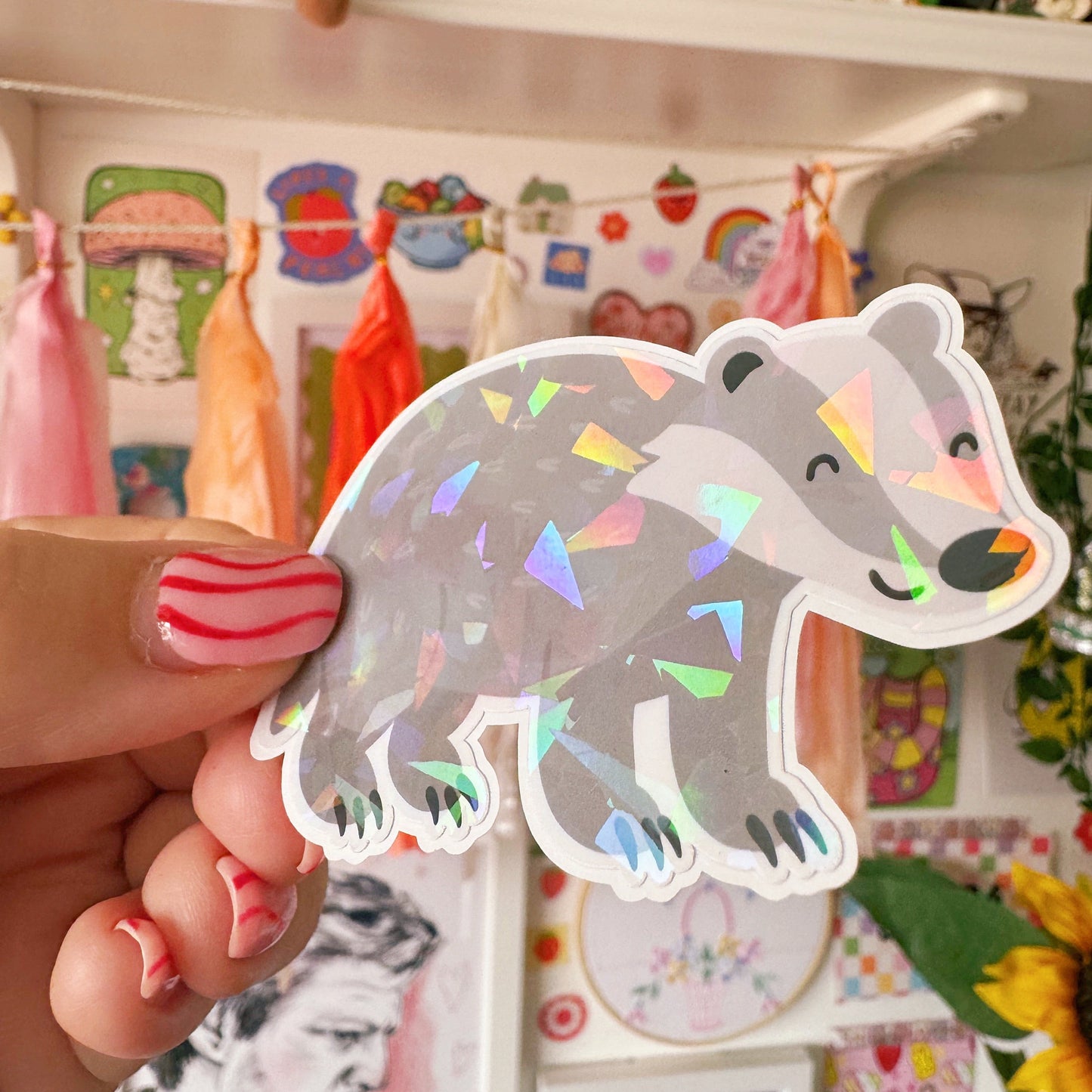 Hand holding a cute badger sticker in front of a shelf with toys and decorations.