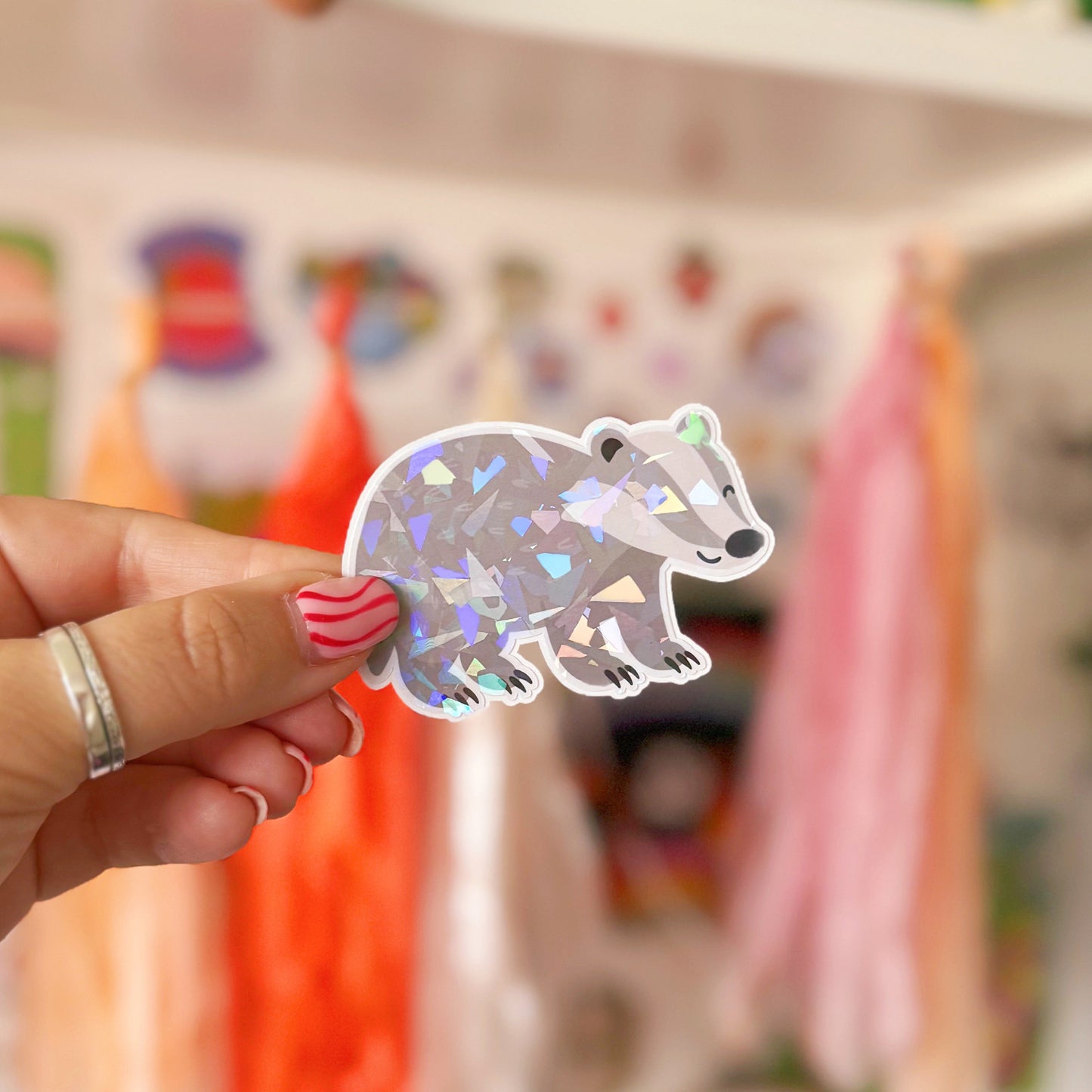 Hand holding a cute badger sticker in front of a shelf with toys and decorations.