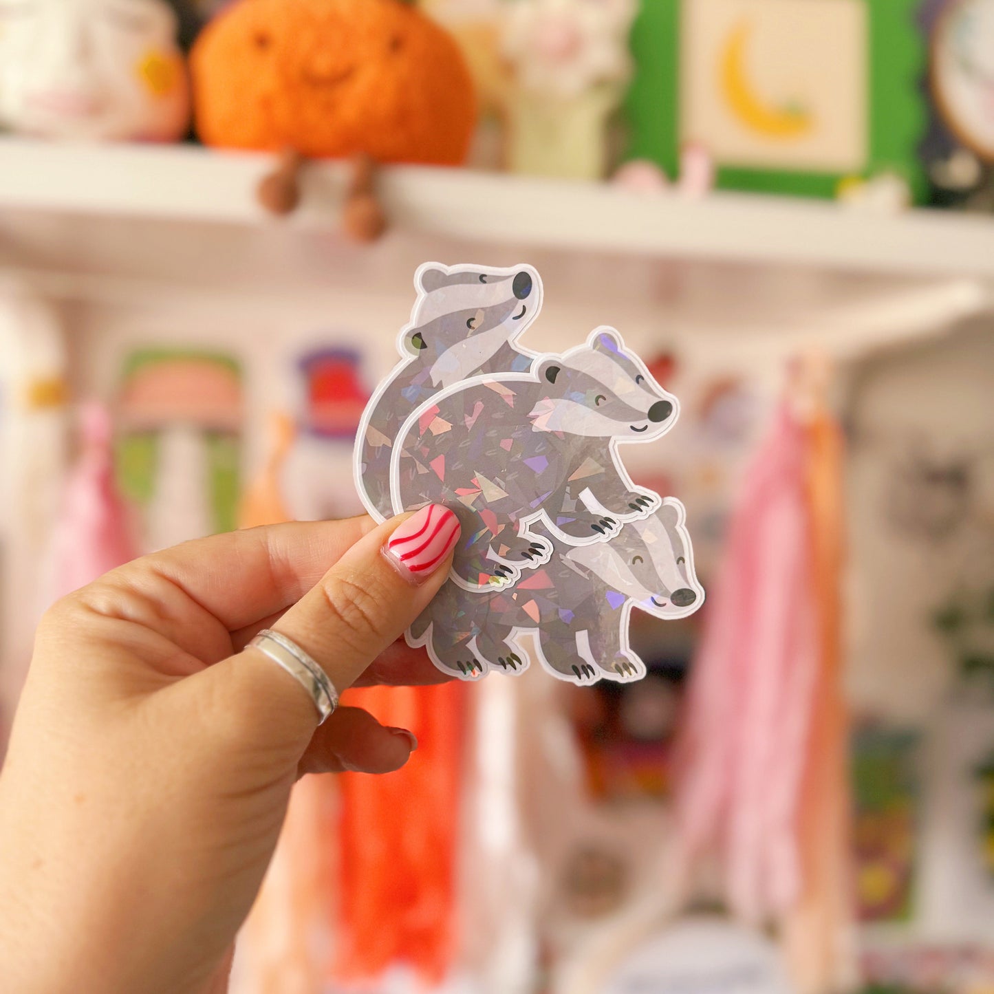 Hand holding a cute badger sticker in front of a shelf with toys and decorations.