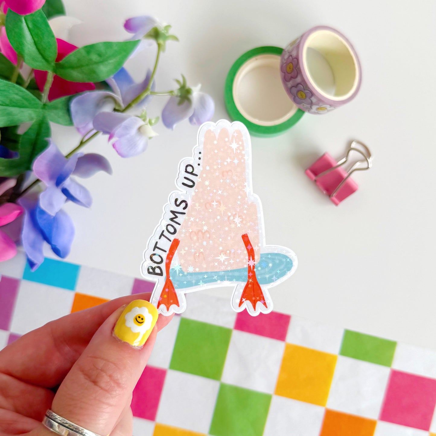 A holographic sticker of a duck upside down in water, with the text - bottoms up, next to it.