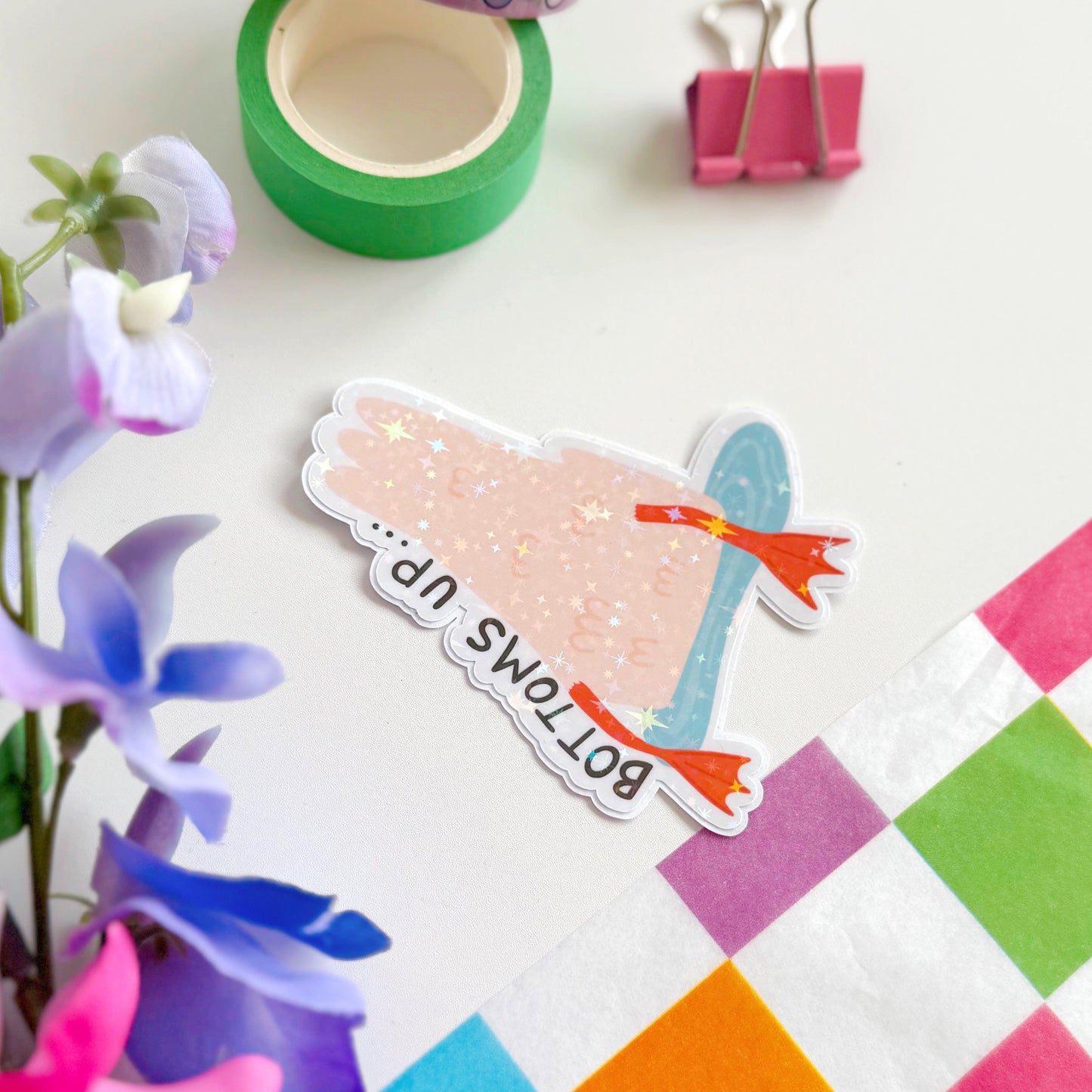 A holographic sticker of a duck upside down in water, with the text - bottoms up, next to it.