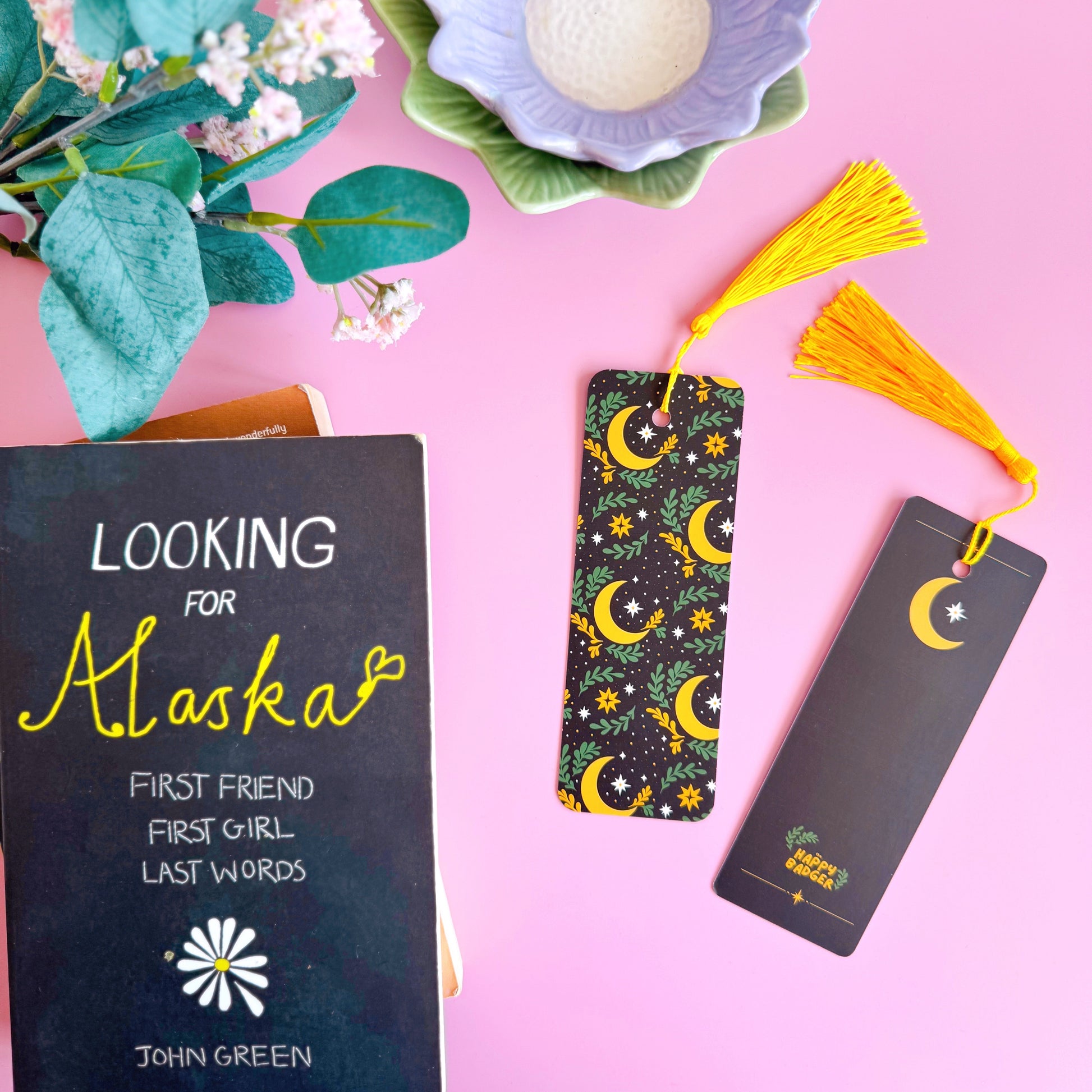 A black bookmark, with a yellow and green celestial themed pattern. It features large moons, green fern leaves and little yellow and white stars. The bookmark has a coordinating tassel in yellow.