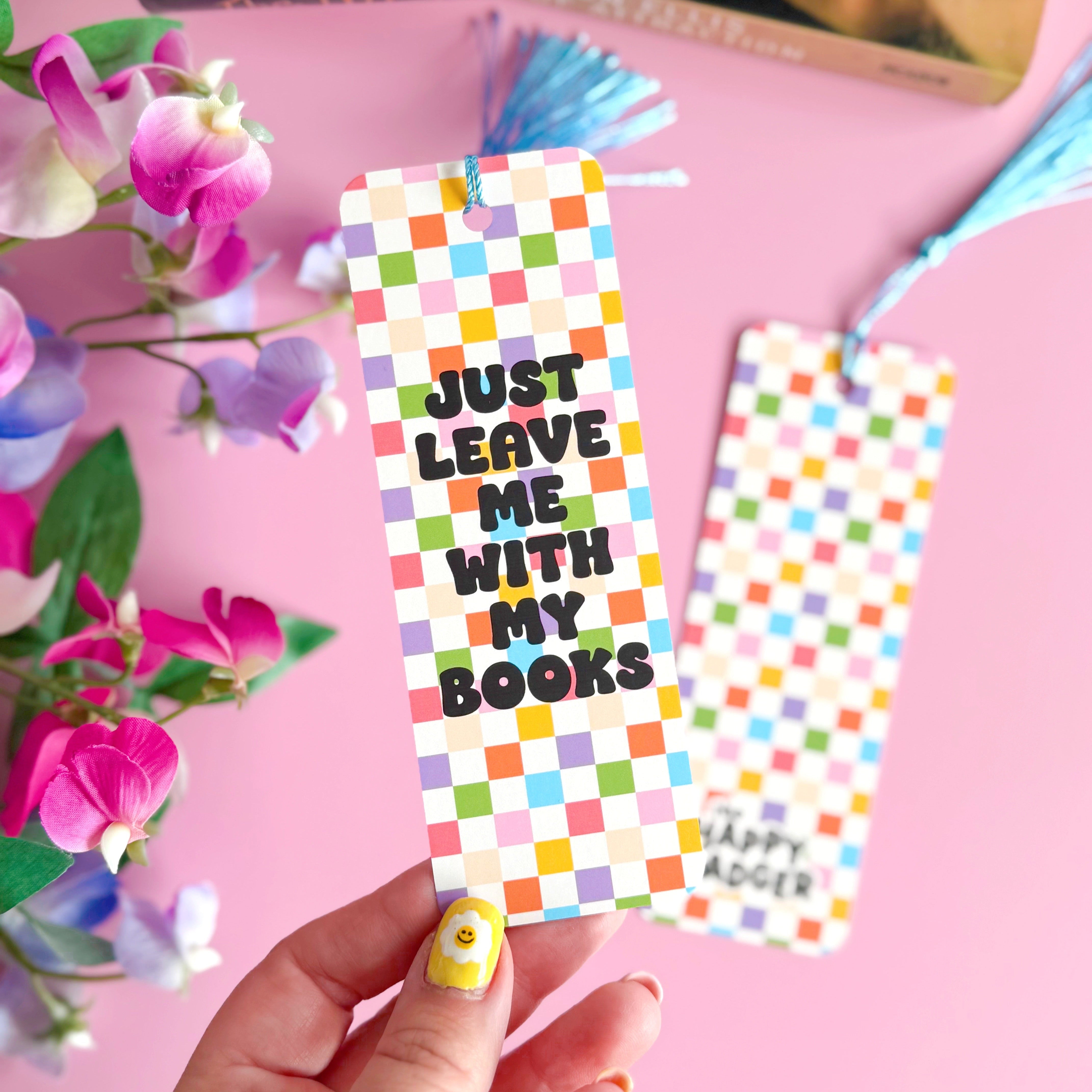 A checkerboard bookmark with rounded corners and a blue tassel, with the words 'just leave me with my books' in bold black writing on the front, and a branding logo on the back.