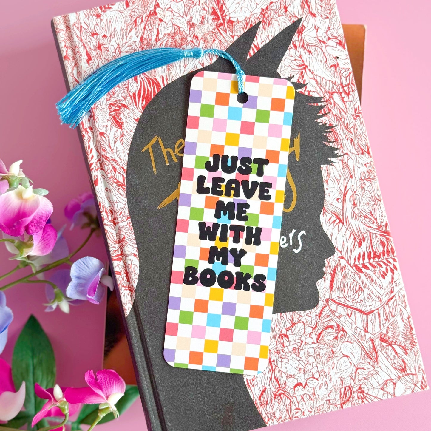 A checkerboard bookmark with rounded corners and a blue tassel, with the words 'just leave me with my books' in bold black writing on the front, and a branding logo on the back.
