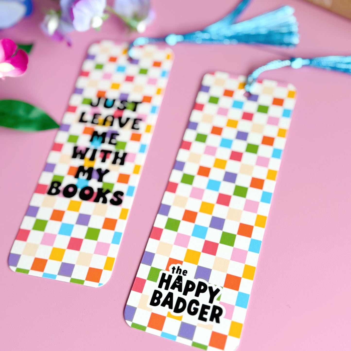 A checkerboard bookmark with rounded corners and a blue tassel, with the words 'just leave me with my books' in bold black writing on the front, and a branding logo on the back.