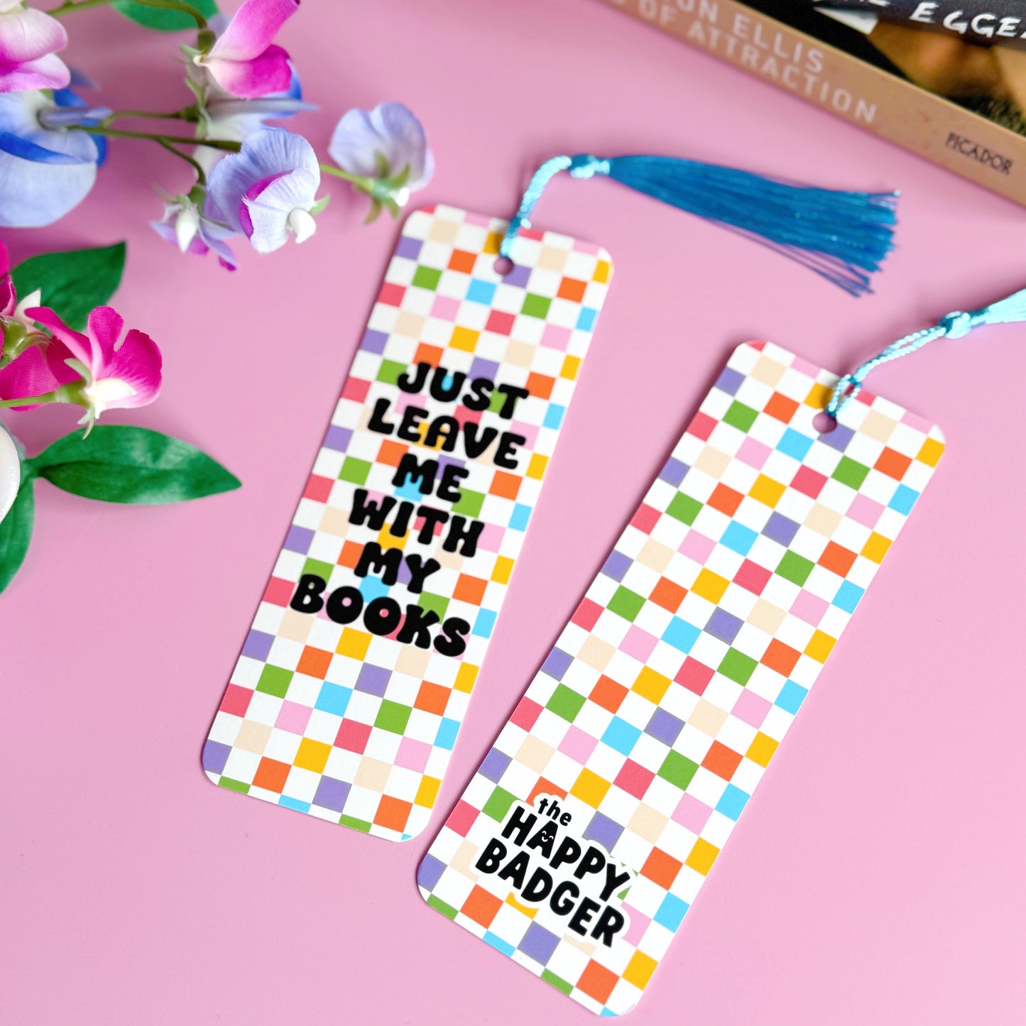 A checkerboard bookmark with rounded corners and a blue tassel, with the words 'just leave me with my books' in bold black writing on the front, and a branding logo on the back.
