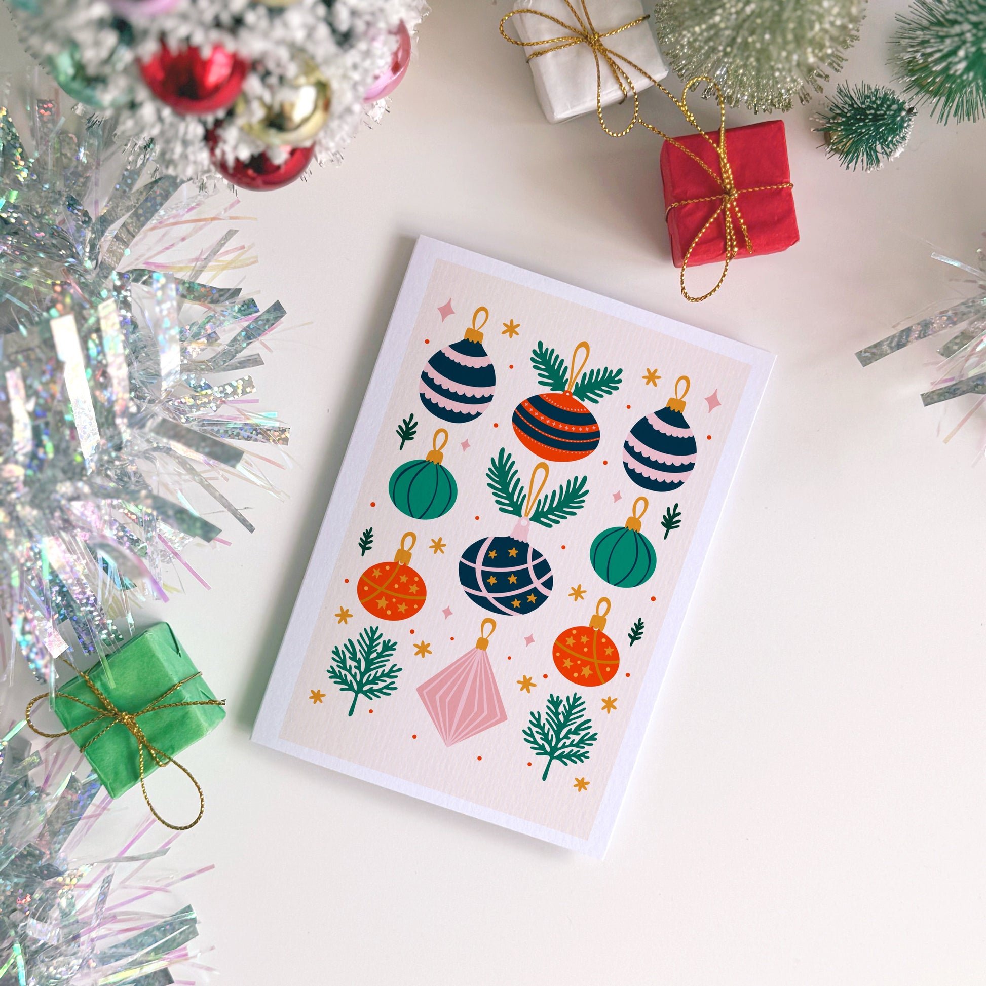 Christmas card with colorful ornaments on a festive background with tinsel and small trees.