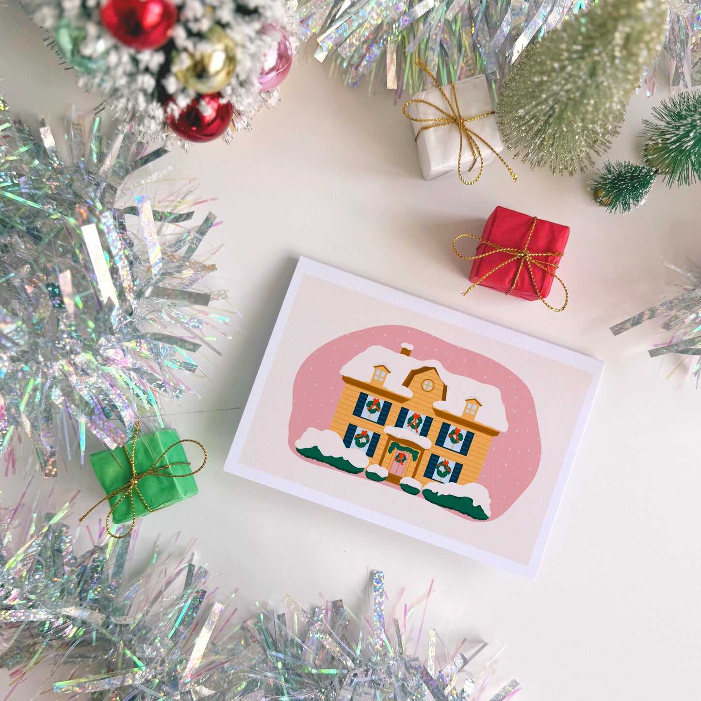 Christmas card with a house illustration surrounded by tinsel and small ornaments on a white surface.