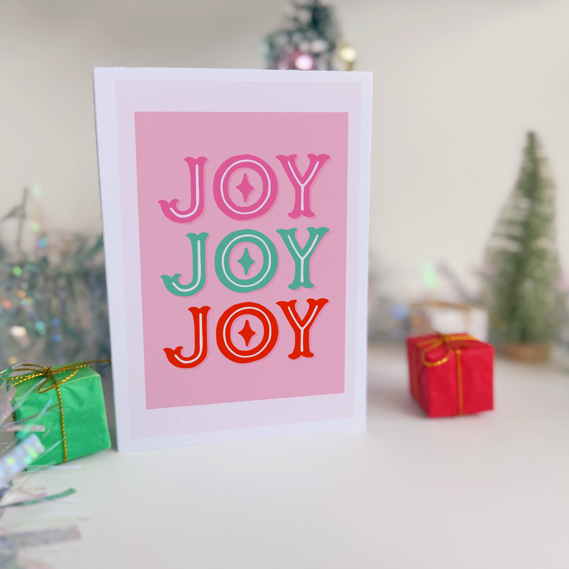 Card with 'JOY JOY JOY' text in colorful letters on a pink background, with Christmas decorations in the background.