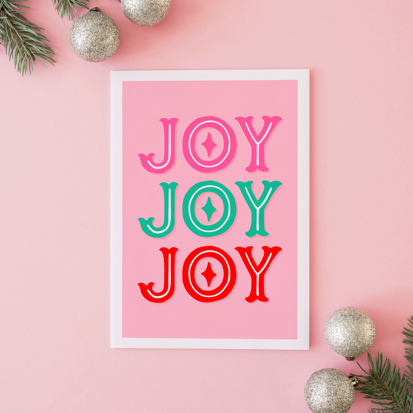 Framed 'JOY' print on a pink background with Christmas decorations.