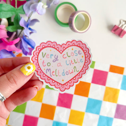 Heart-shaped sticker with text held by a hand, colorful paper, and flowers in the background