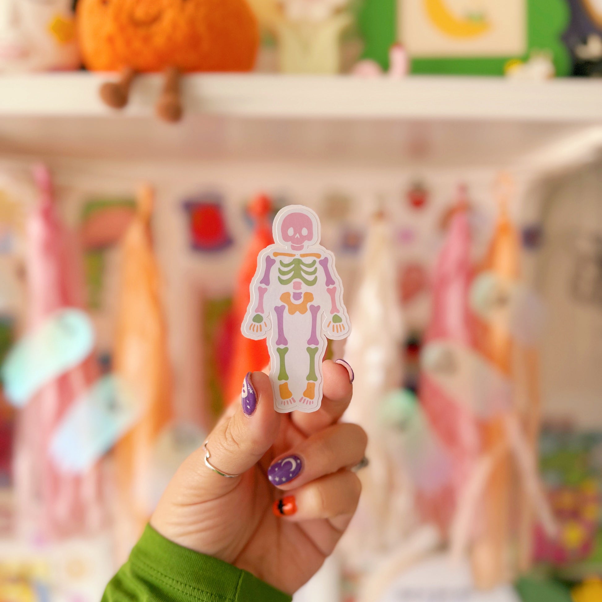 Hand holding a colourful skeleton sticker in front of a blurred toy store background