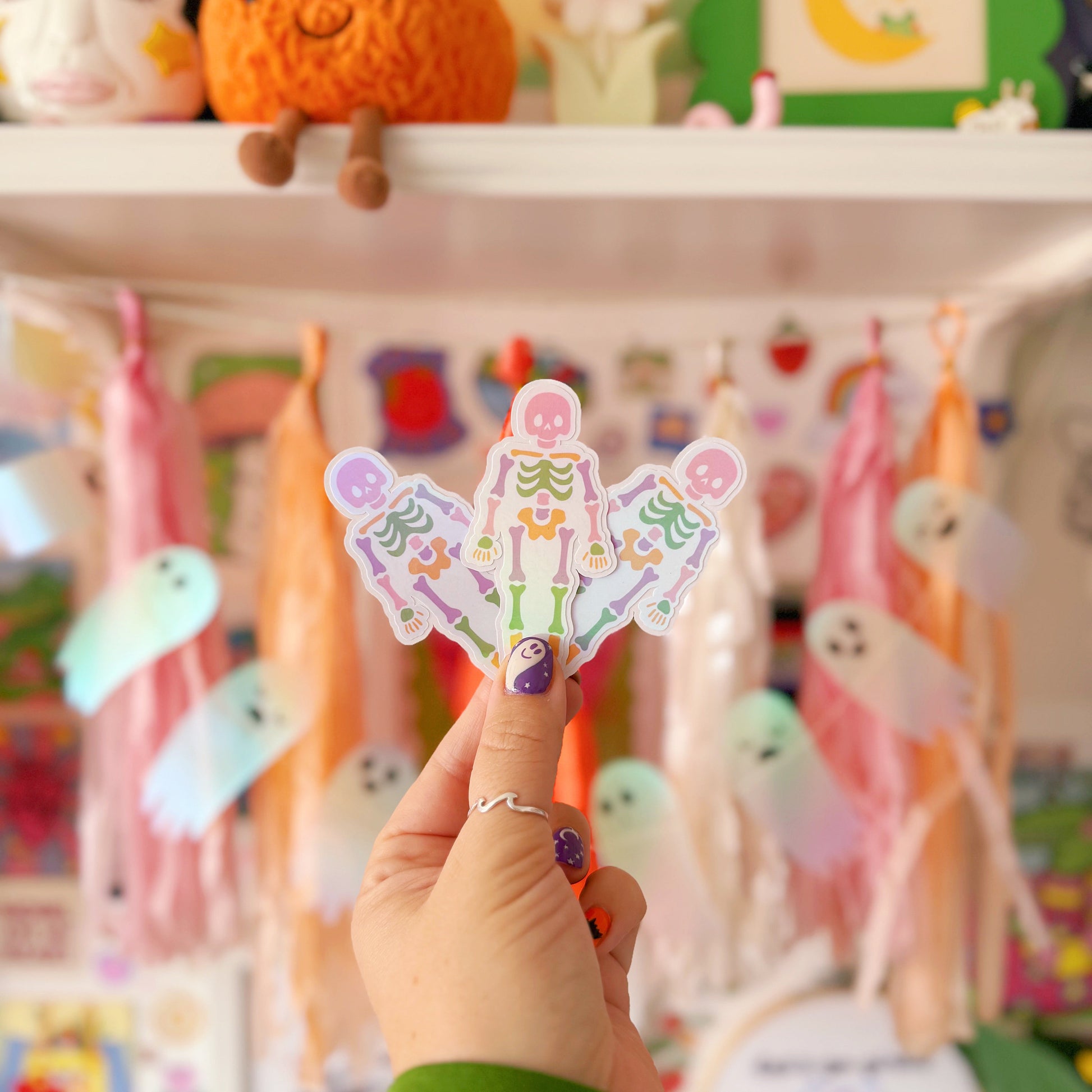 Hand holding 3 colourful skeleton stickers with a decorated background featuring toys and decorations.