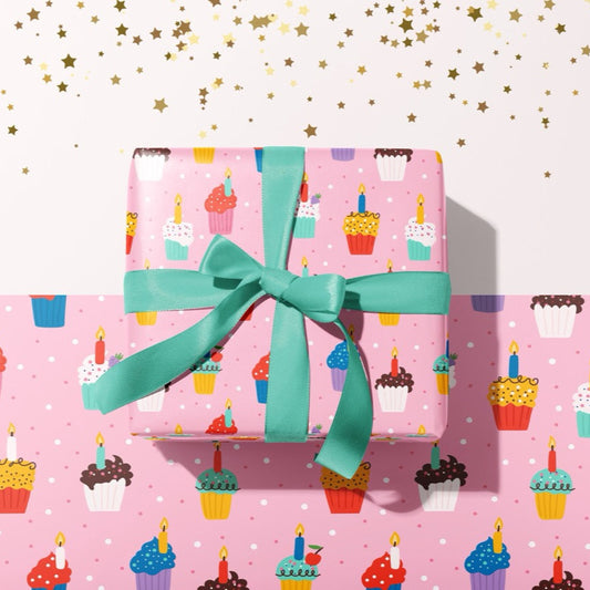A present wrapped in pink gift wrap, which has colourful cupcakes and candles all over. The present is wrapped with a teal bow.