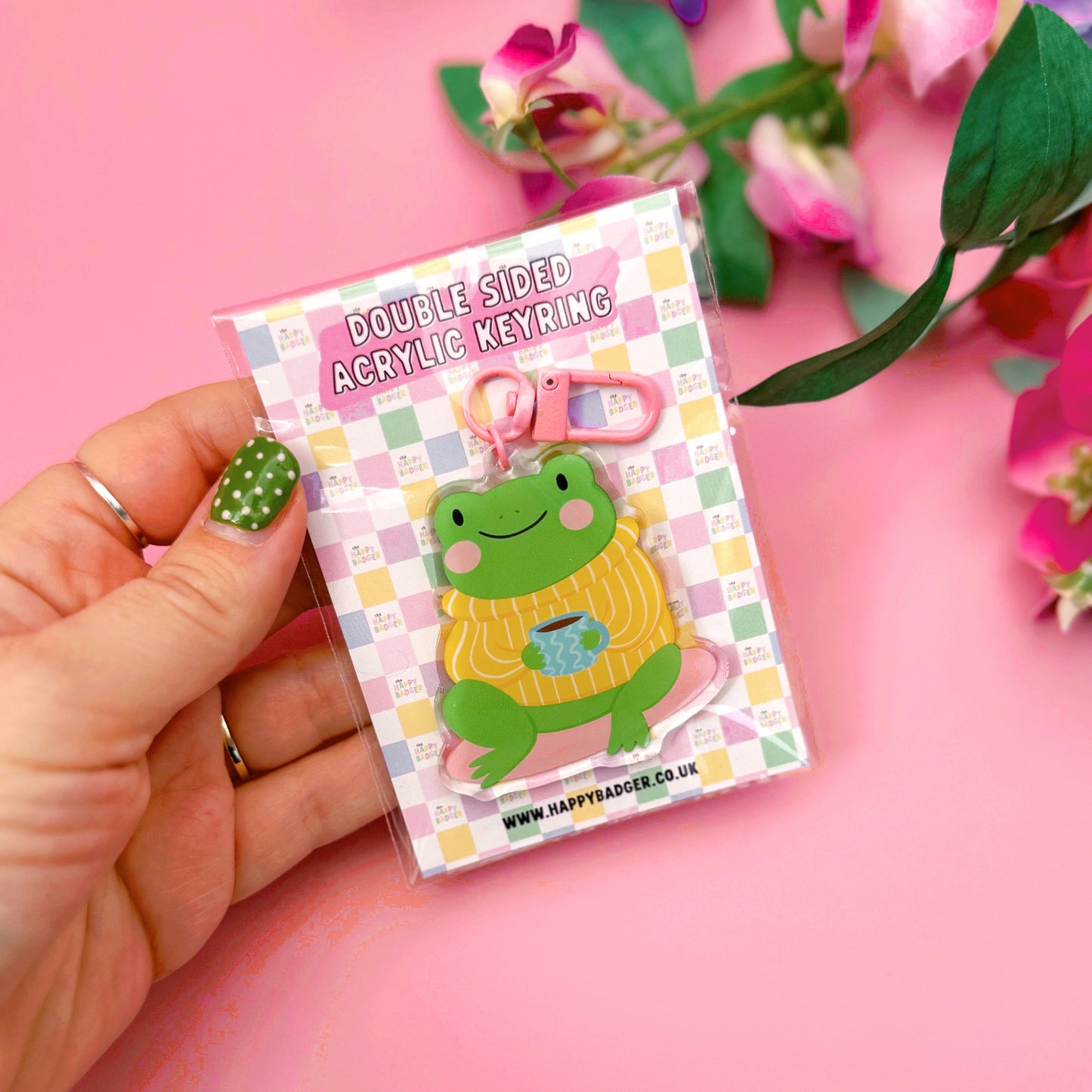 Hand holding a keyring with a frog design on a pink background with flowers.