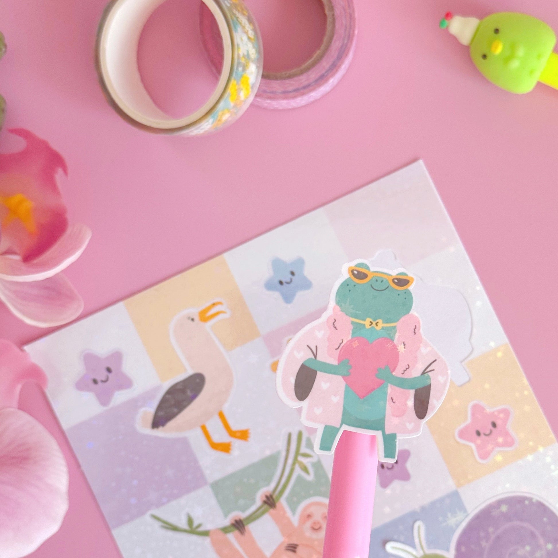 A sticker from the mini sticker pals sticker sheet. This is a frog holding a heart, coated with a holographic laminate with little starbursts on.