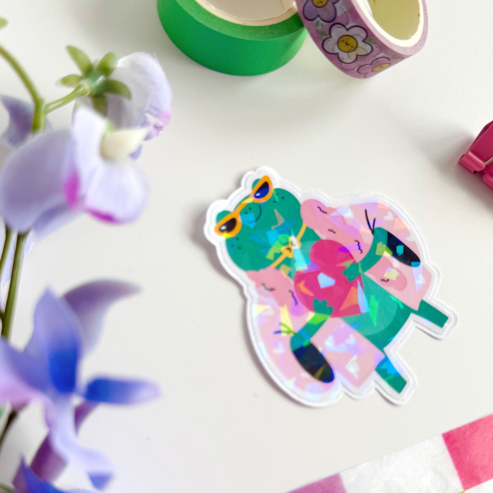A holographic sticker, of a green frog with a pink coat on holding a love heart. The sticker has an easy peel lip.