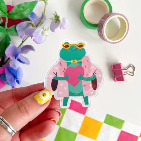 A holographic sticker, of a green frog with a pink coat on holding a love heart. The sticker has an easy peel lip.