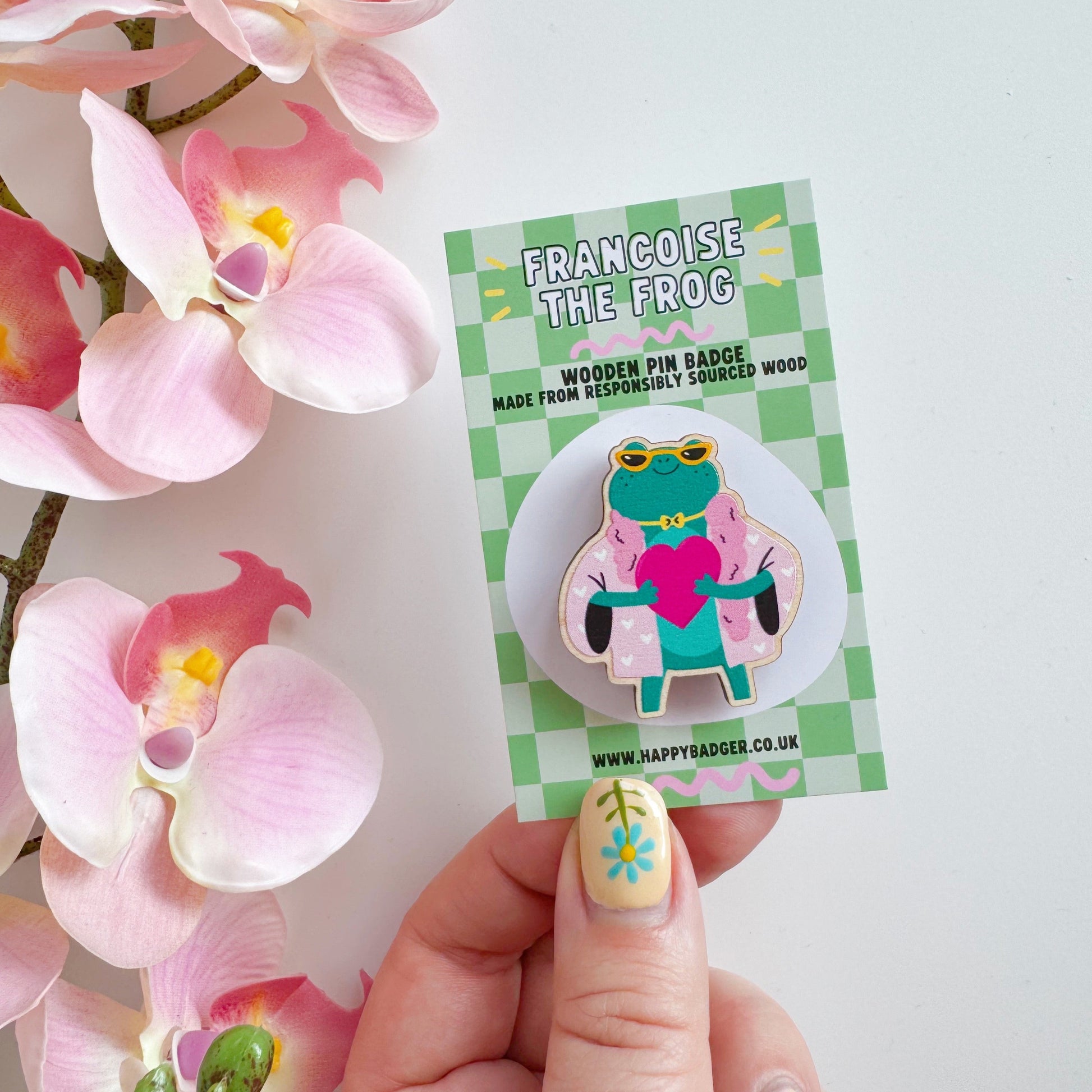 A cute illustration of a frog in sunglasses and a pink coat, holding a love heart, on an eco friendly wooden pin badge with two back fastenings.