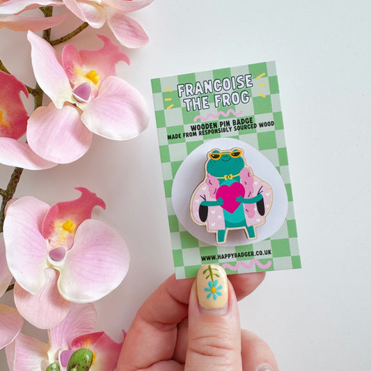 A cute illustration of a frog in sunglasses and a pink coat, holding a love heart, on an eco friendly wooden pin badge with two back fastenings.