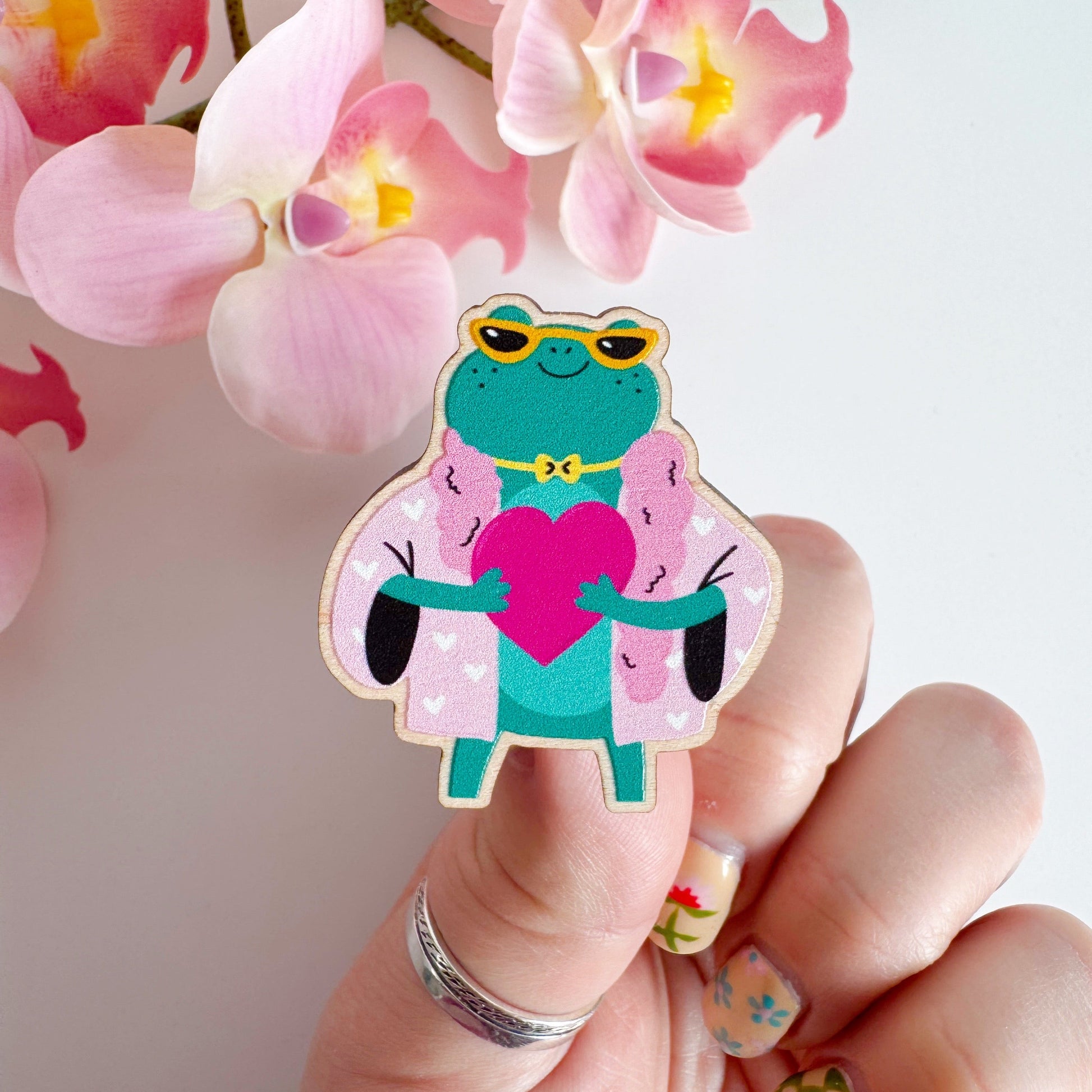 A cute illustration of a frog in sunglasses and a pink coat, holding a love heart, on an eco friendly wooden pin badge with two back fastenings.