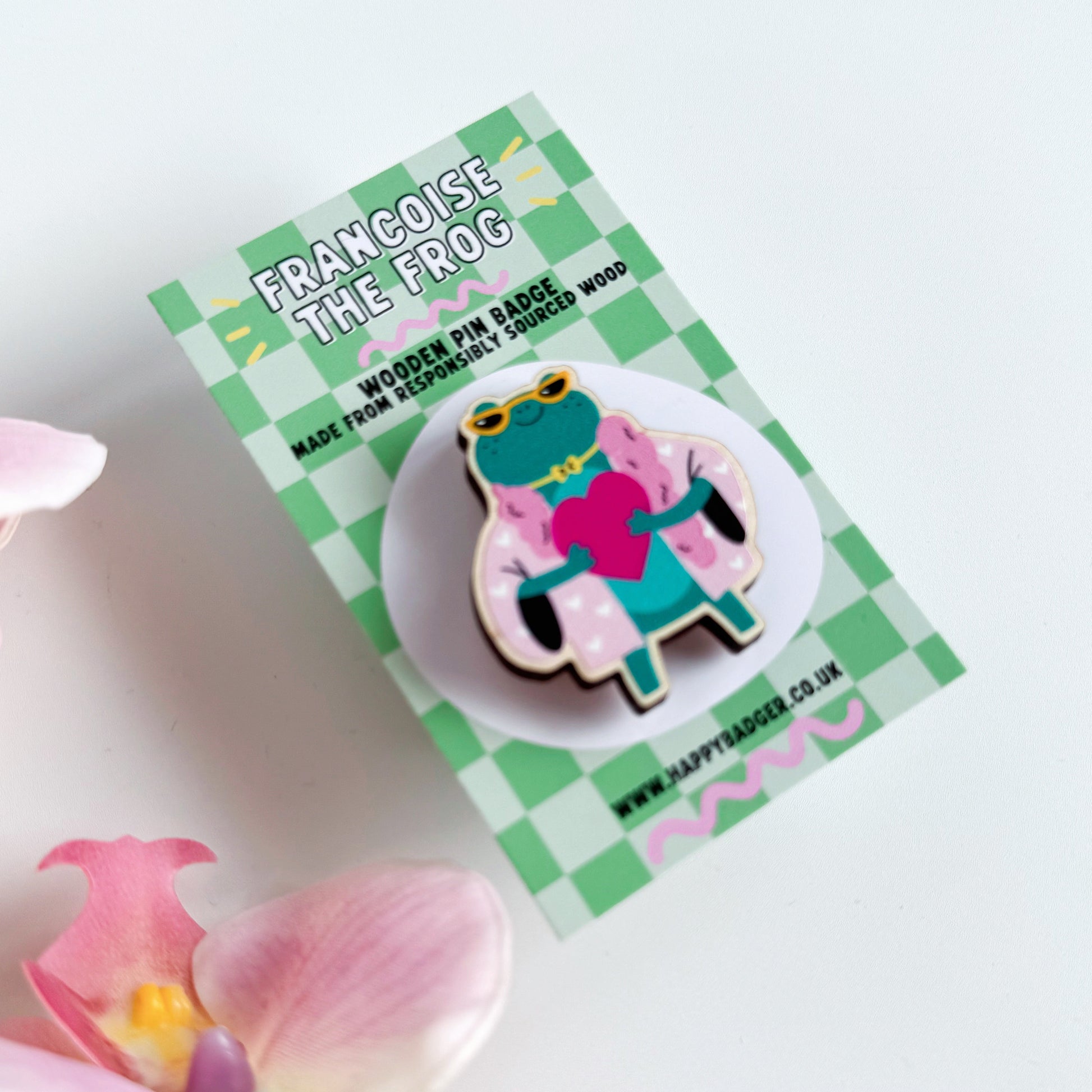 A cute illustration of a frog in sunglasses and a pink coat, holding a love heart, on an eco friendly wooden pin badge with two back fastenings.