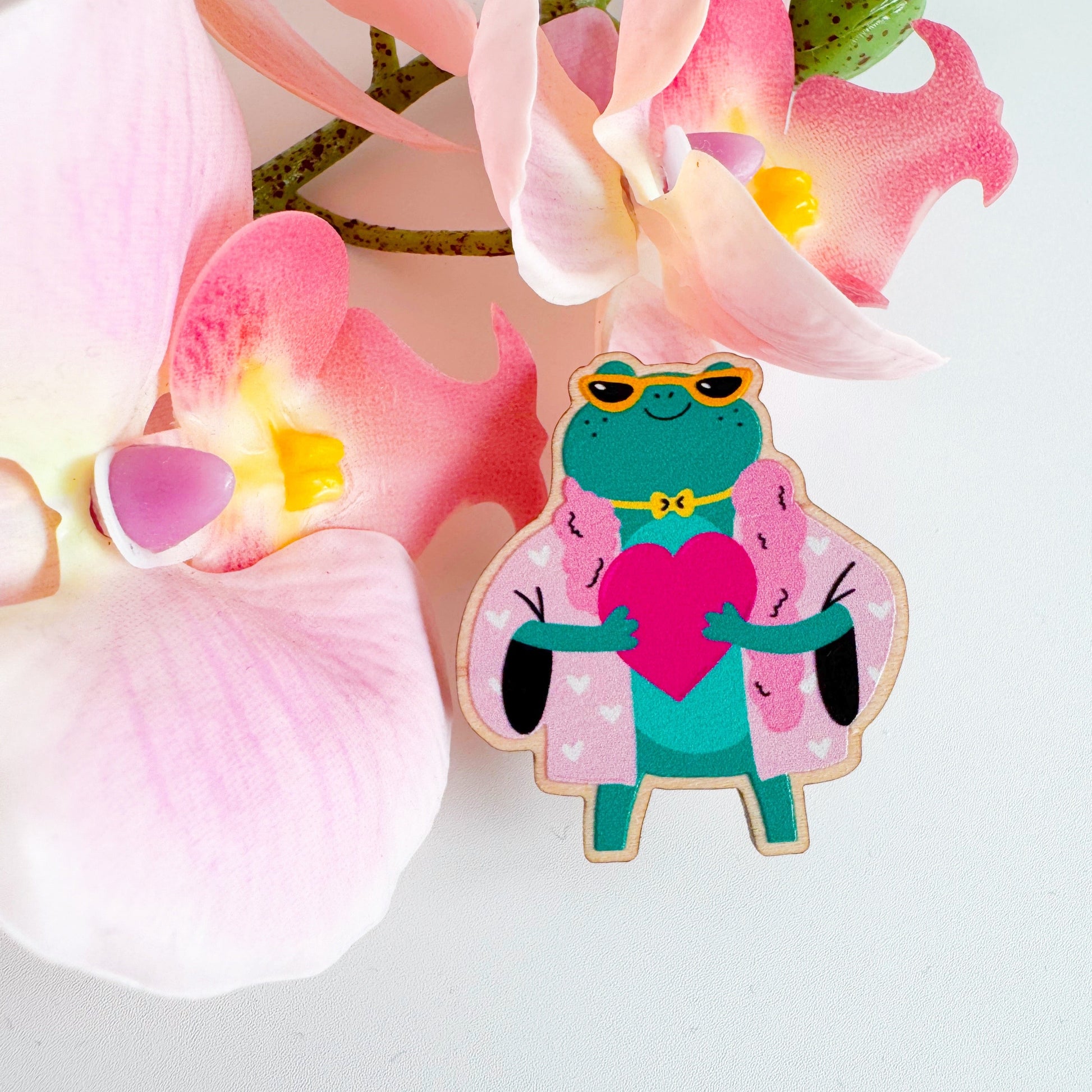 A cute illustration of a frog in sunglasses and a pink coat, holding a love heart, on an eco friendly wooden pin badge with two back fastenings.
