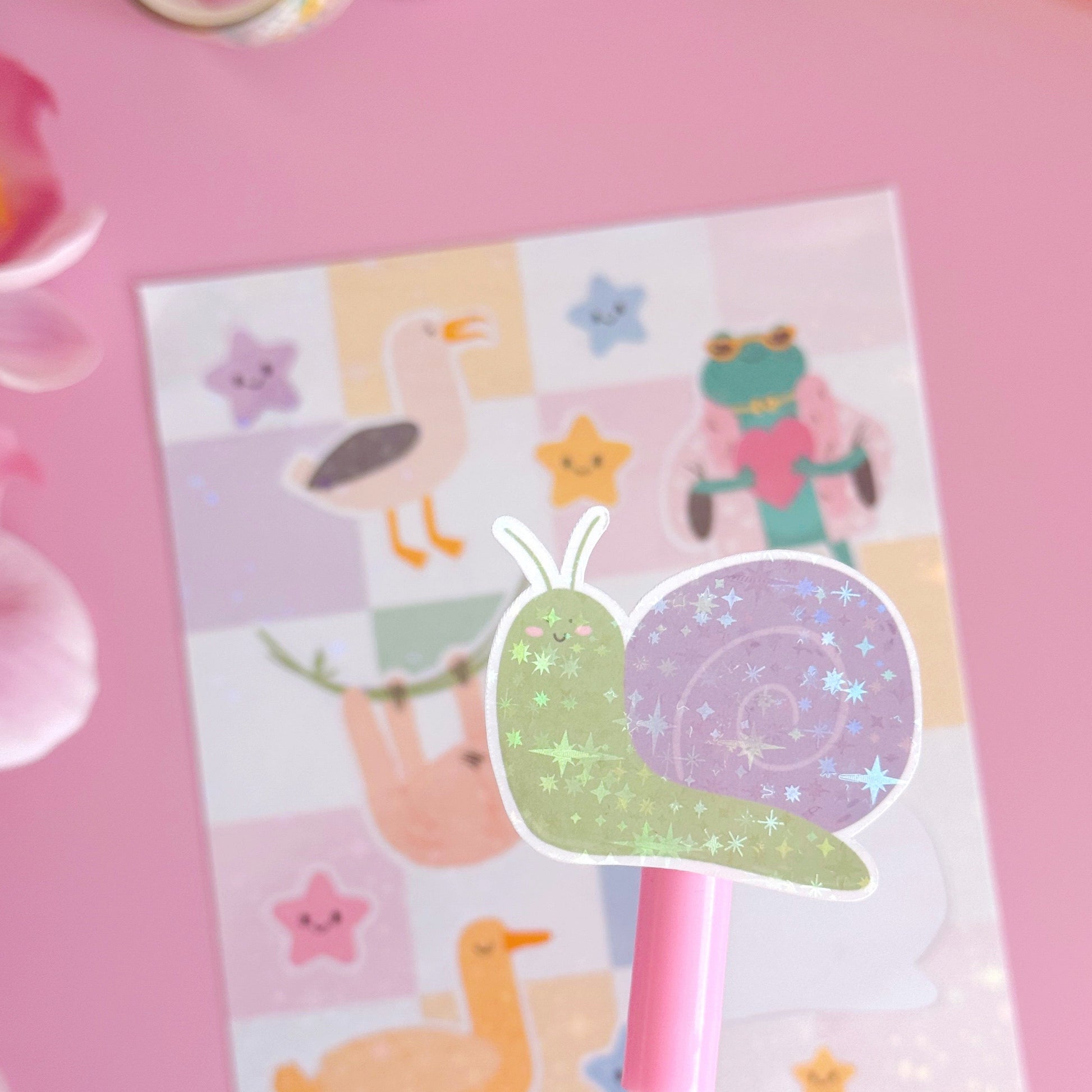 A sticker from the mini sticker pals sticker sheet. This is a purple and green snail, coated with a holographic laminate with little starbursts on.