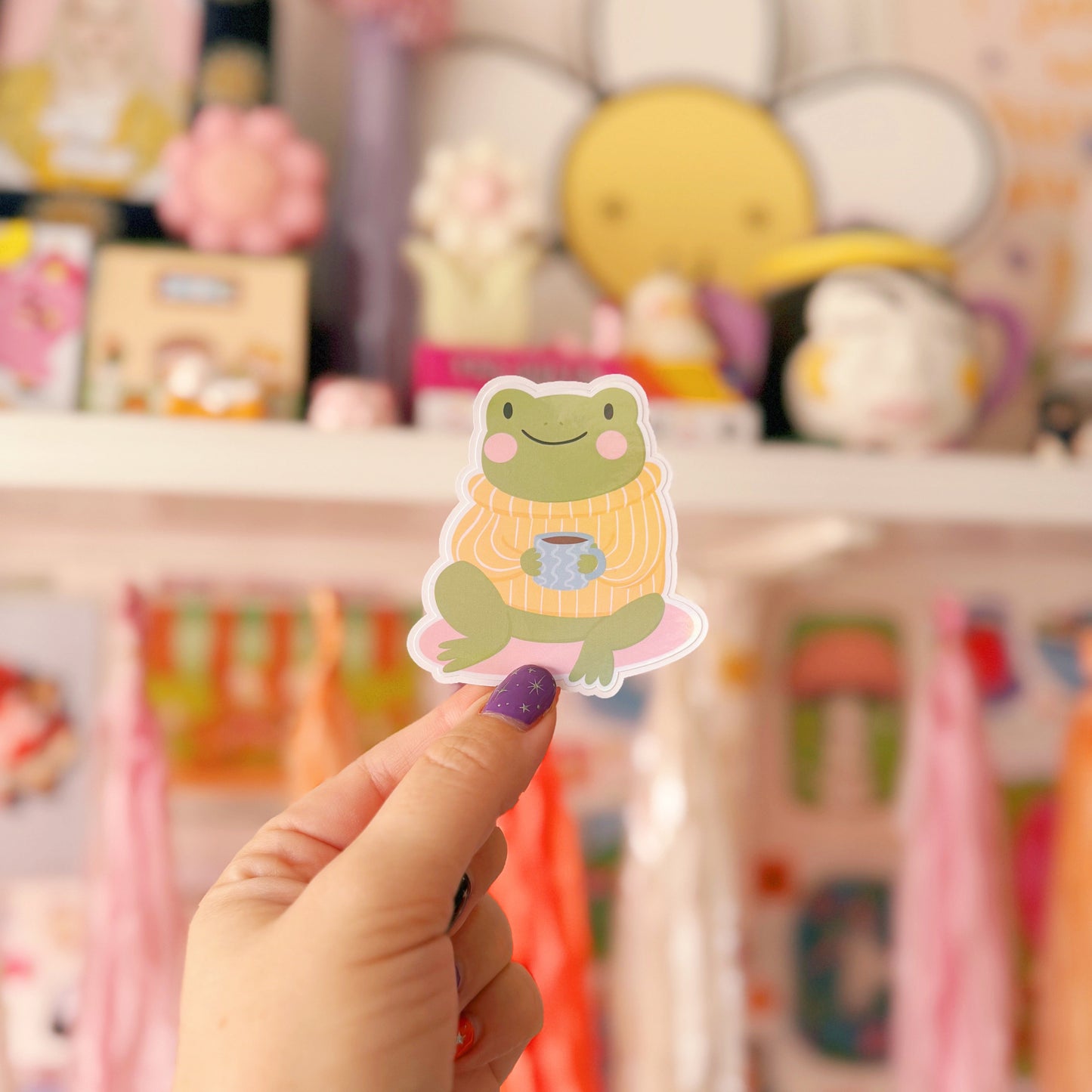 A sticker of a cute little frog, sitting down wearing a yellow striped jumper. The frog is also holding a mug of coffee/tea.