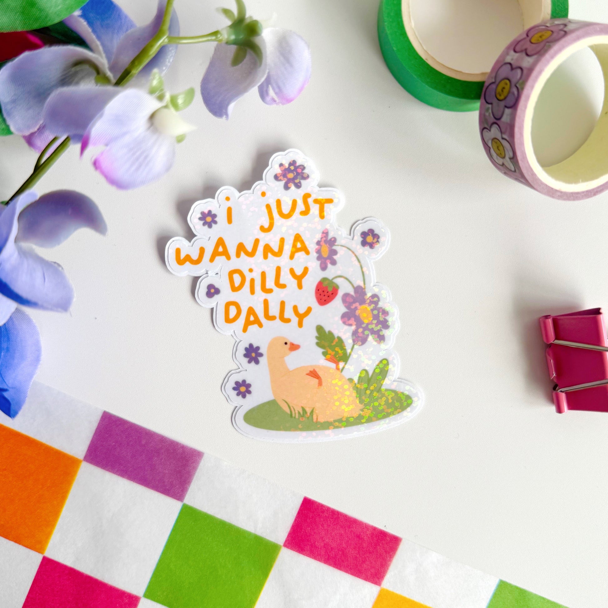 A cute holographic sticker with a duck laying on it's back, next to purple flowers. Above it are the words 'I Just Wanna Dilly Dally' in yellow.