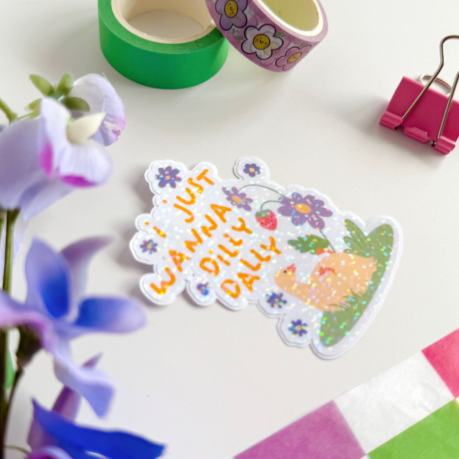 A cute holographic sticker with a duck laying on it's back, next to purple flowers. Above it are the words 'I Just Wanna Dilly Dally' in yellow.