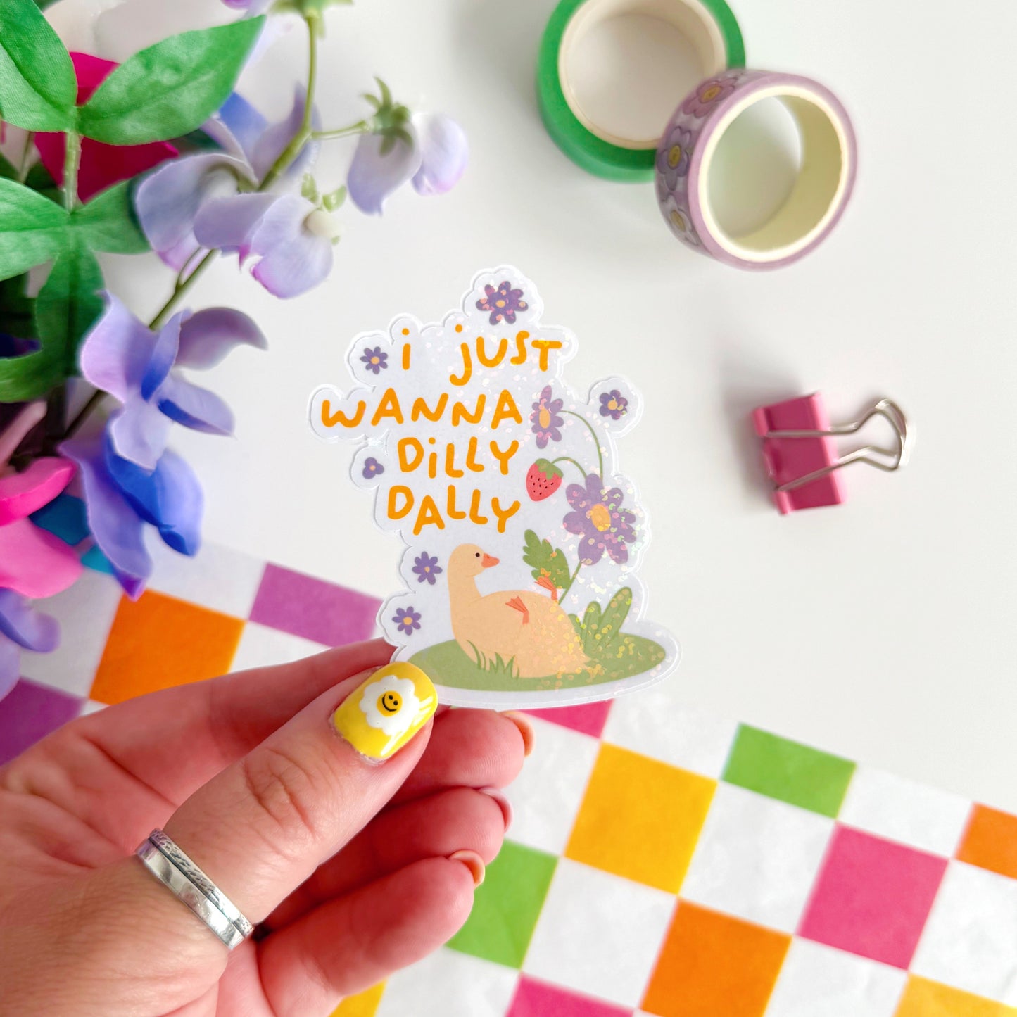 A cute holographic sticker with a duck laying on it's back, next to purple flowers. Above it are the words 'I Just Wanna Dilly Dally' in yellow.
