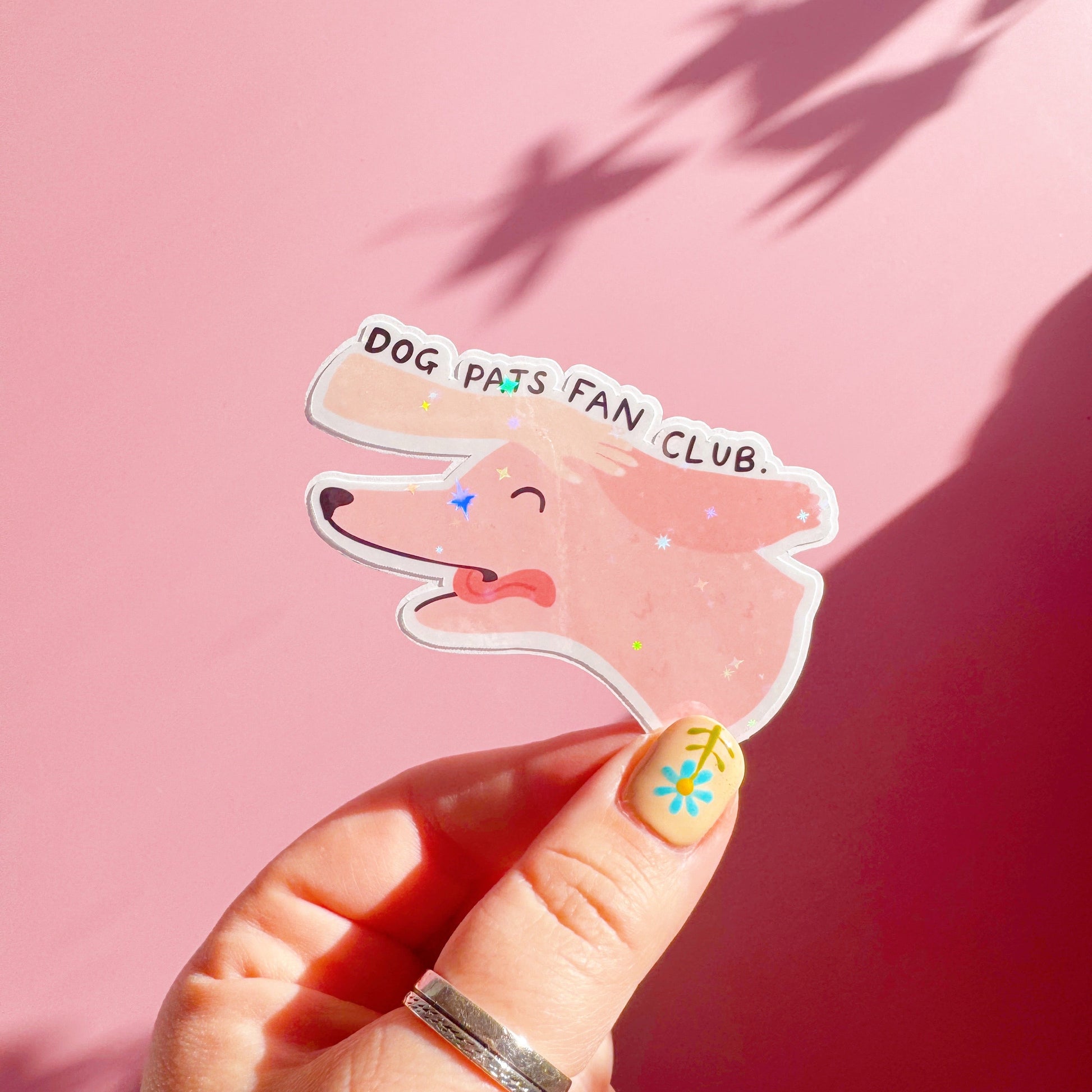 A holographic sticker of a pink dog, with its tongue sticking out, with a hand patting its head. The text reads 'Dog Pats Fan Club.'