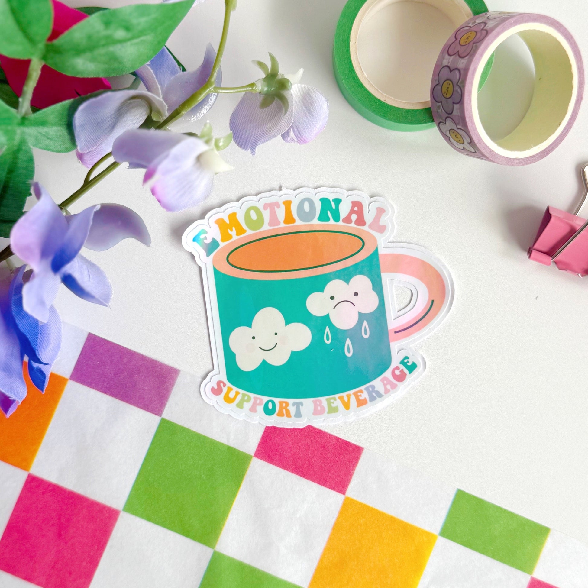 A sticker with an illustration of a mug - that has a happy cloud and a sad cloud on the front, with the words 'emotional support beverage' in colourful lettering around the mug.