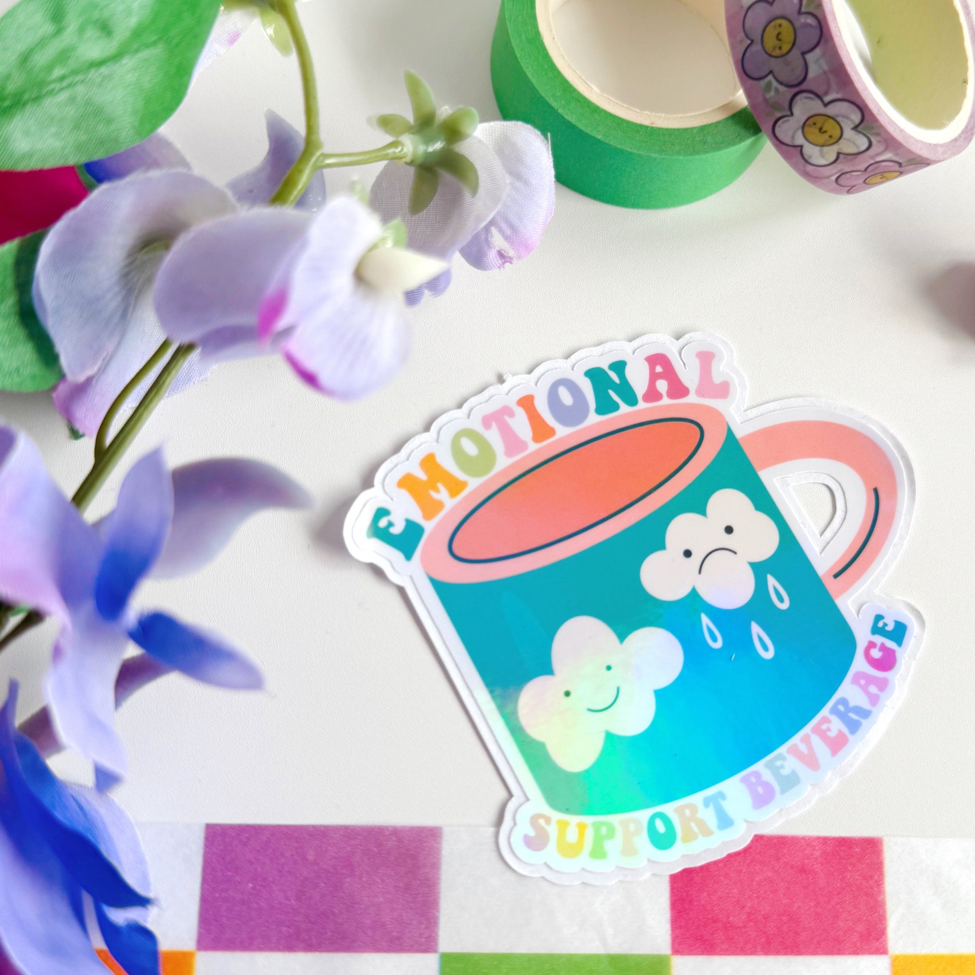 A sticker with an illustration of a mug - that has a happy cloud and a sad cloud on the front, with the words 'emotional support beverage' in colourful lettering around the mug.