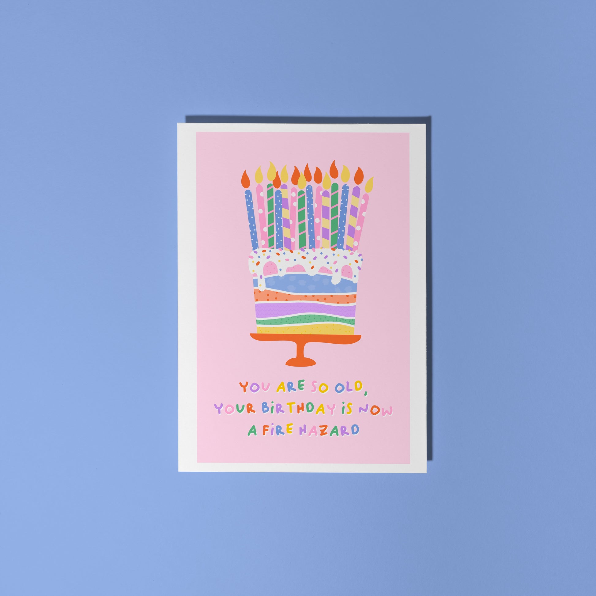 A pink card, with an illustration of a colourful cake on - which has several candles on the top. Underneath in colourful text, it says 'you are so old, your birthday is now a fire hazard'