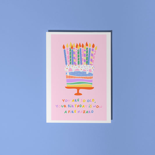 A pink card, with an illustration of a colourful cake on - which has several candles on the top. Underneath in colourful text, it says 'you are so old, your birthday is now a fire hazard'