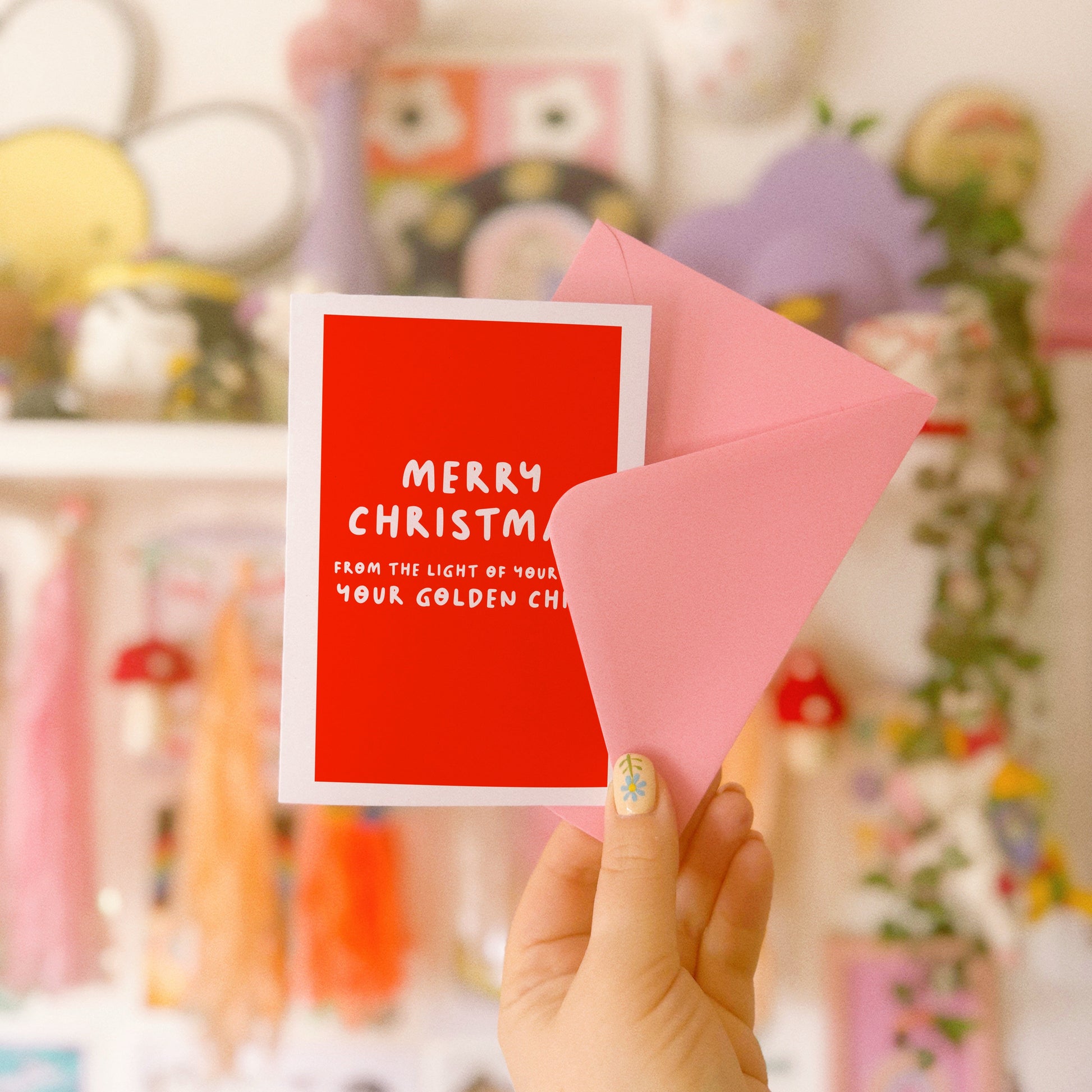 Hand holding a red Christmas card with a pink envelope against a colourful background.