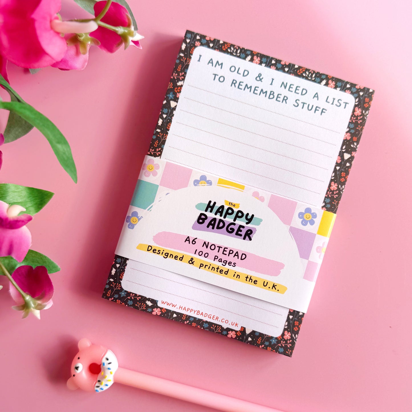 Notepad with floral design and 'Happy Badger' branding on a pink background with flowers and a pen.