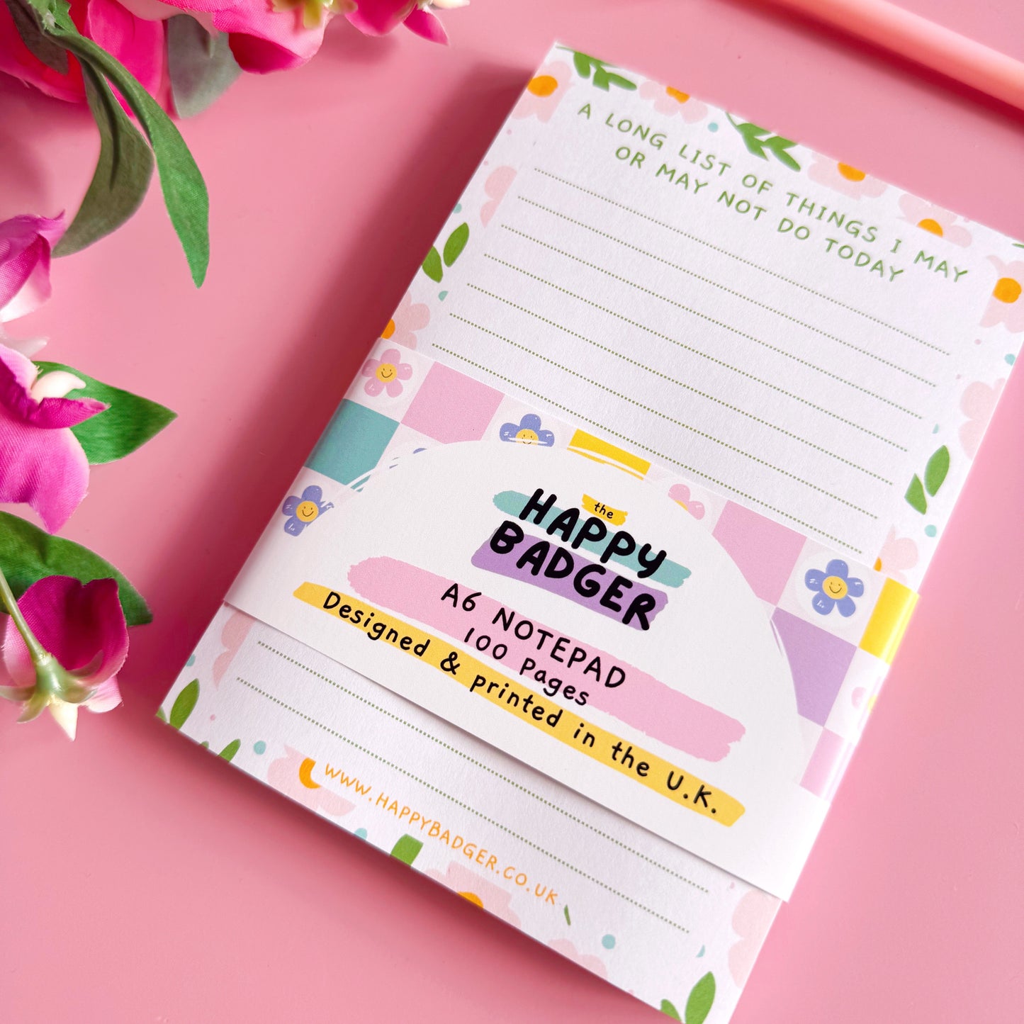Notepad with floral design and 'Happy Badger' branding on a pink background with flowers and a pen.