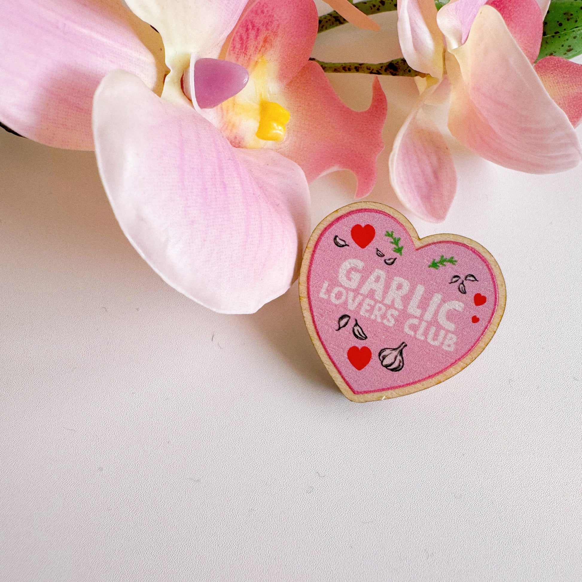 A pink heart shaped wooden pin badge, with the words 'garlic lovers club' on the front