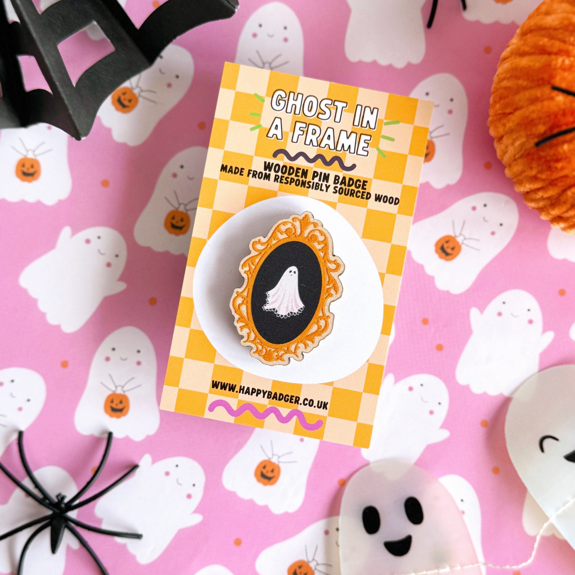 A wooden eco friendly pin badge of a cute spooky ghost on a black background, in an orange ornate frame.