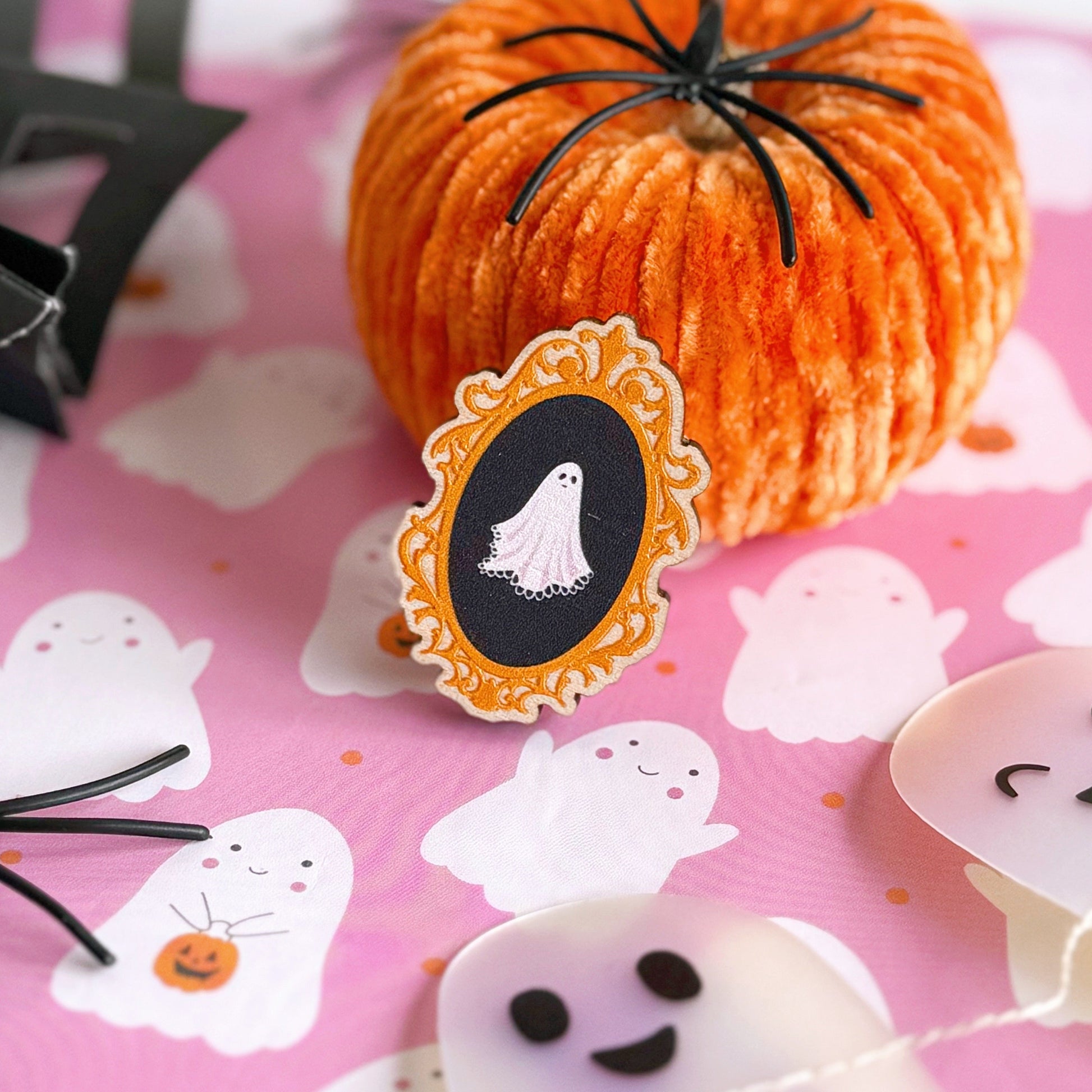 A wooden eco friendly pin badge of a cute spooky ghost on a black background, in an orange ornate frame.