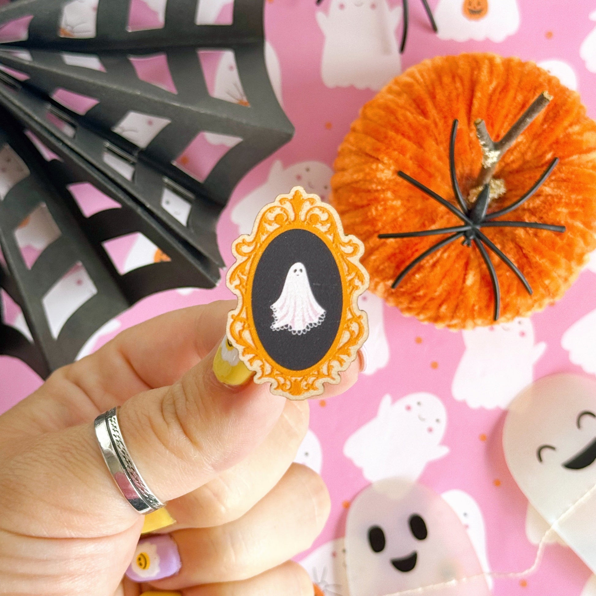 A wooden eco friendly pin badge of a cute spooky ghost on a black background, in an orange ornate frame.